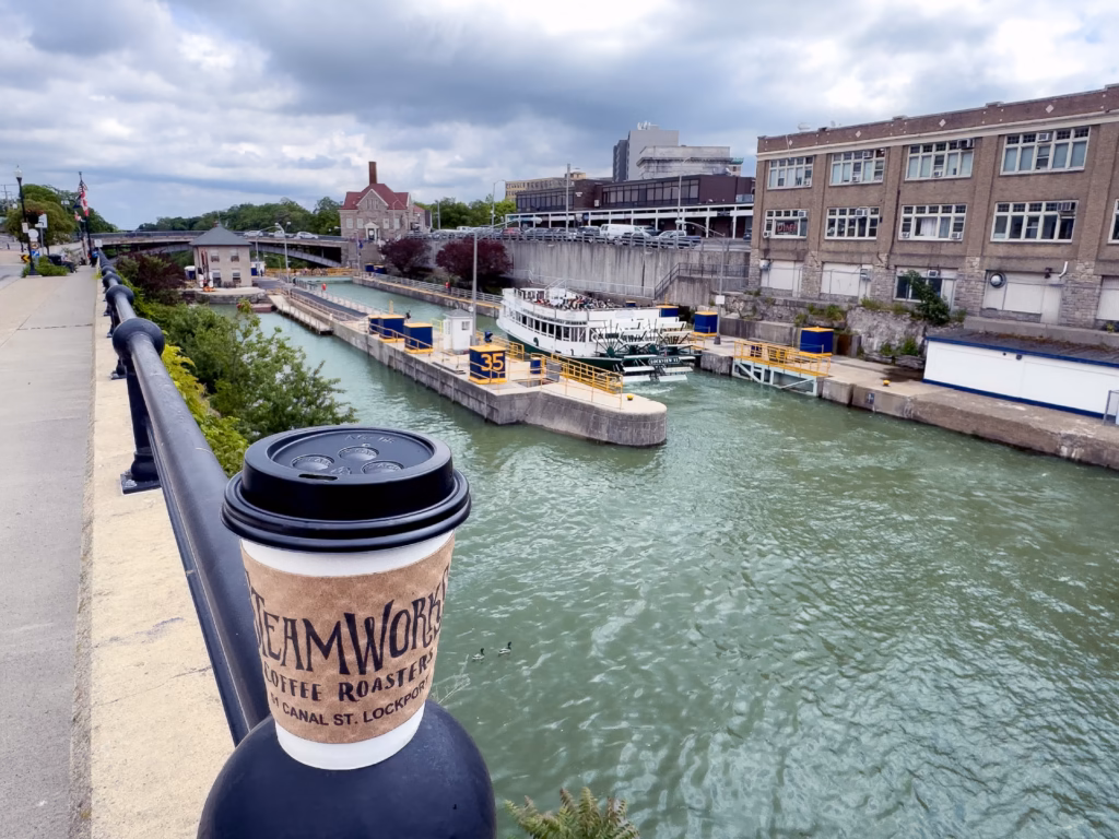 A takeaway cup of Steamworks Coffee Roasters perched on a bridge railing overlooking a boat navigating the historic locks in Lockport, New York, highlighting erie canal specialty coffee experiences.
