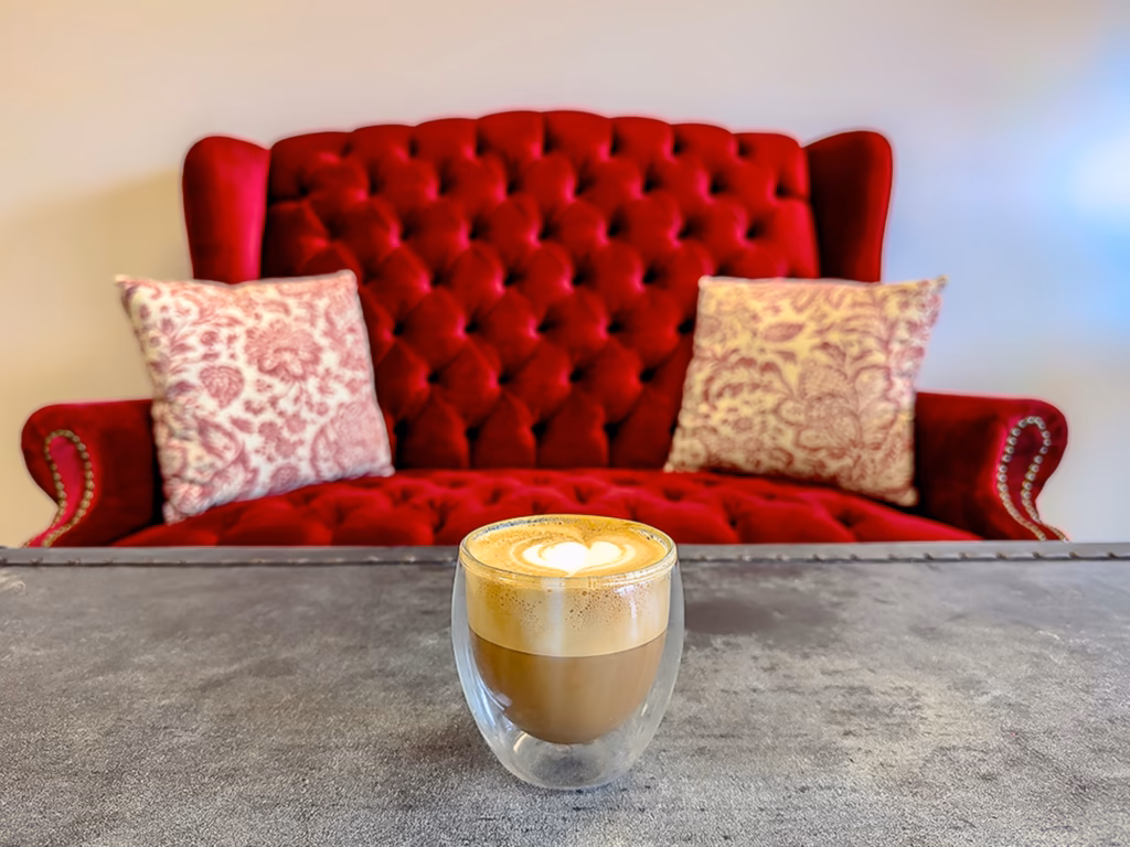A beautifully crafted cappuccino with heart-shaped latte art in a double-walled glass cup, set against a plush red tufted couch at Gypsy Kitchen, a unique erie canal specialty coffee spot in Lockport, New York.