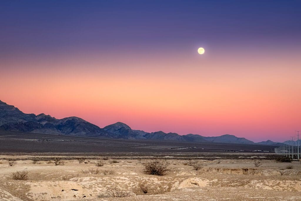 Dusk time in Nevada desert, at the outskirts of town. A full moon rising above a range of mountains in a rugged landscape and endless sky. Soft pastels and fiery hues.