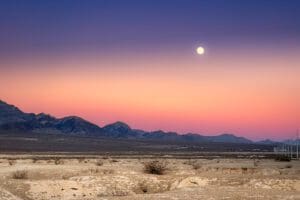 Dusk time in Nevada desert, at the outskirts of Las Vegas. A full moon rising above a range of mountains in a rugged landscape and endless sky. Soft pastels and fiery hues.