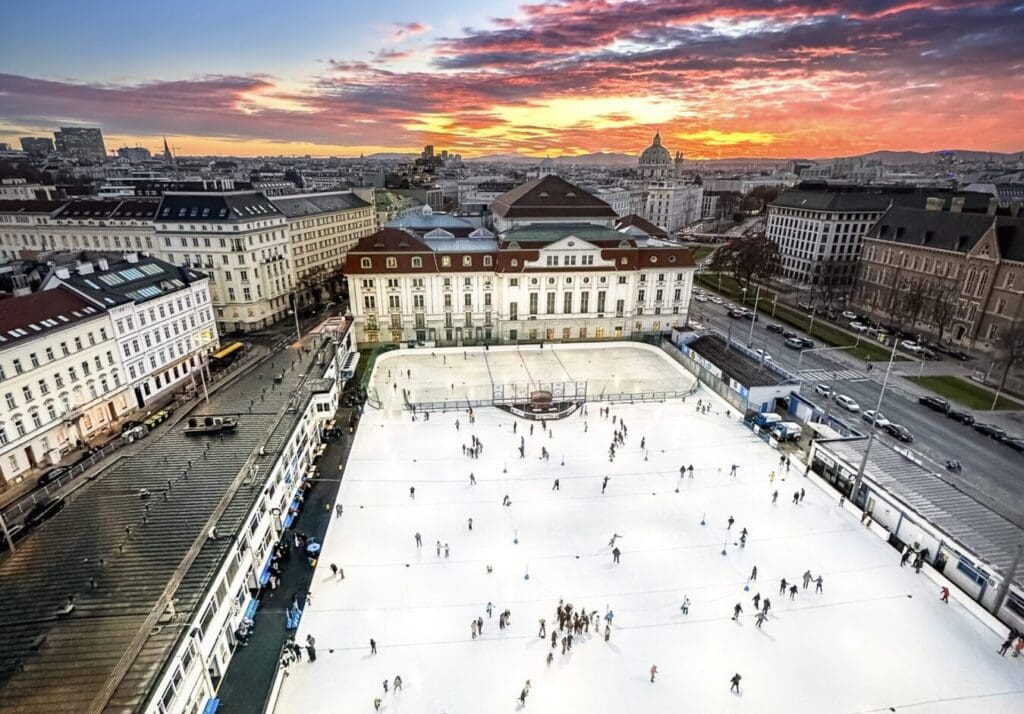 Vienna in the winter, means outdoor sports, like hockey and figure skating. Exterior winter photograph overlooking district 1 of Vienna with an exterior massive ice rink in the foreground, a hockey rink in the mid ground and dramatic cloudy skies during a winter's sunset over Carl's Cathedral
