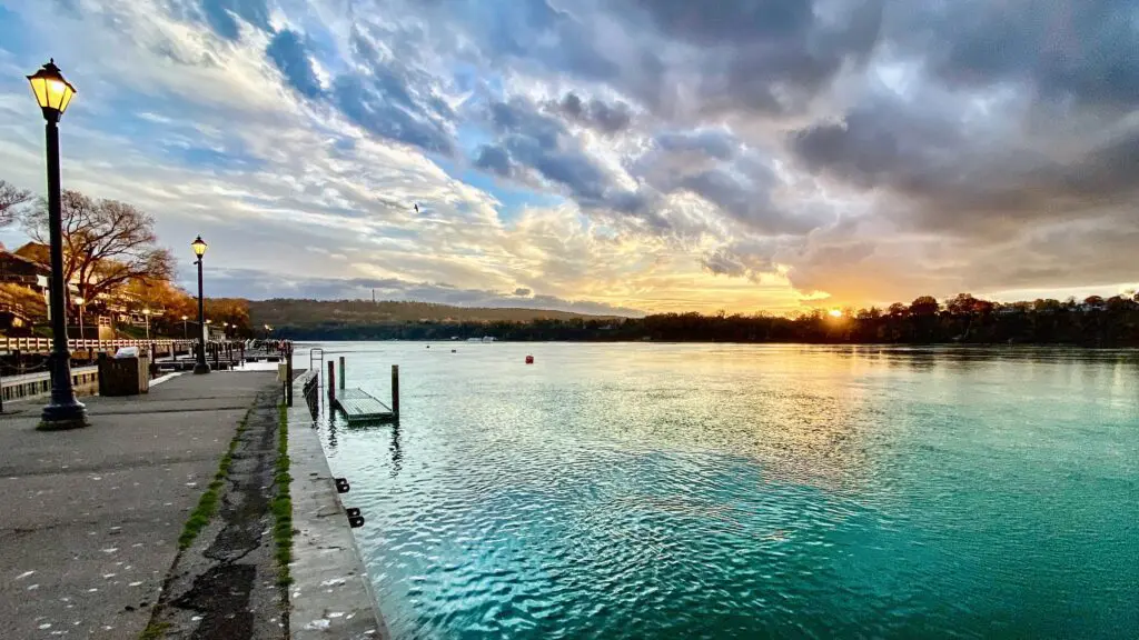 Water landing and boat ramp with promenade highlighted by antique street lamps, while the calm turquoise waters of the Niagara River are gently swaying, while a dramatic cloudy sky is undergoing the changes of colours during sunset.