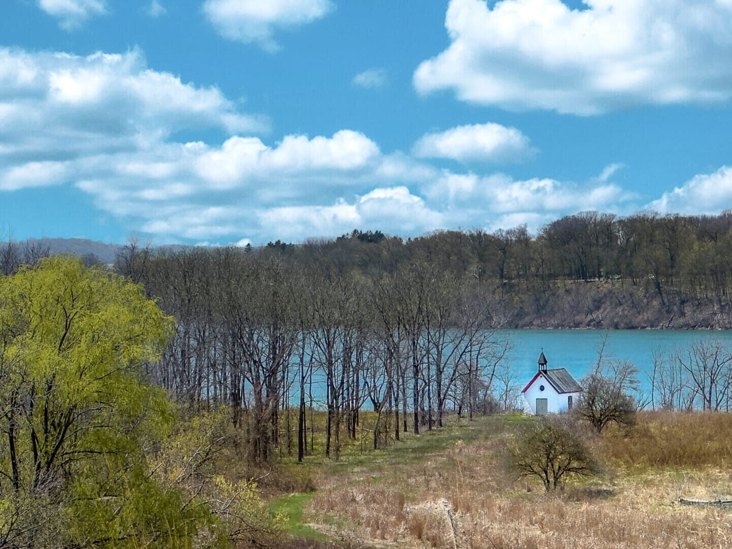 Lewiston, New York Stella Niagara preserve with trees starting to bloom, tall grass covers the foreground in front of a small white isolated church, the turquoise waters of the mighty Niagara River flow past, and a motor boat skims the water