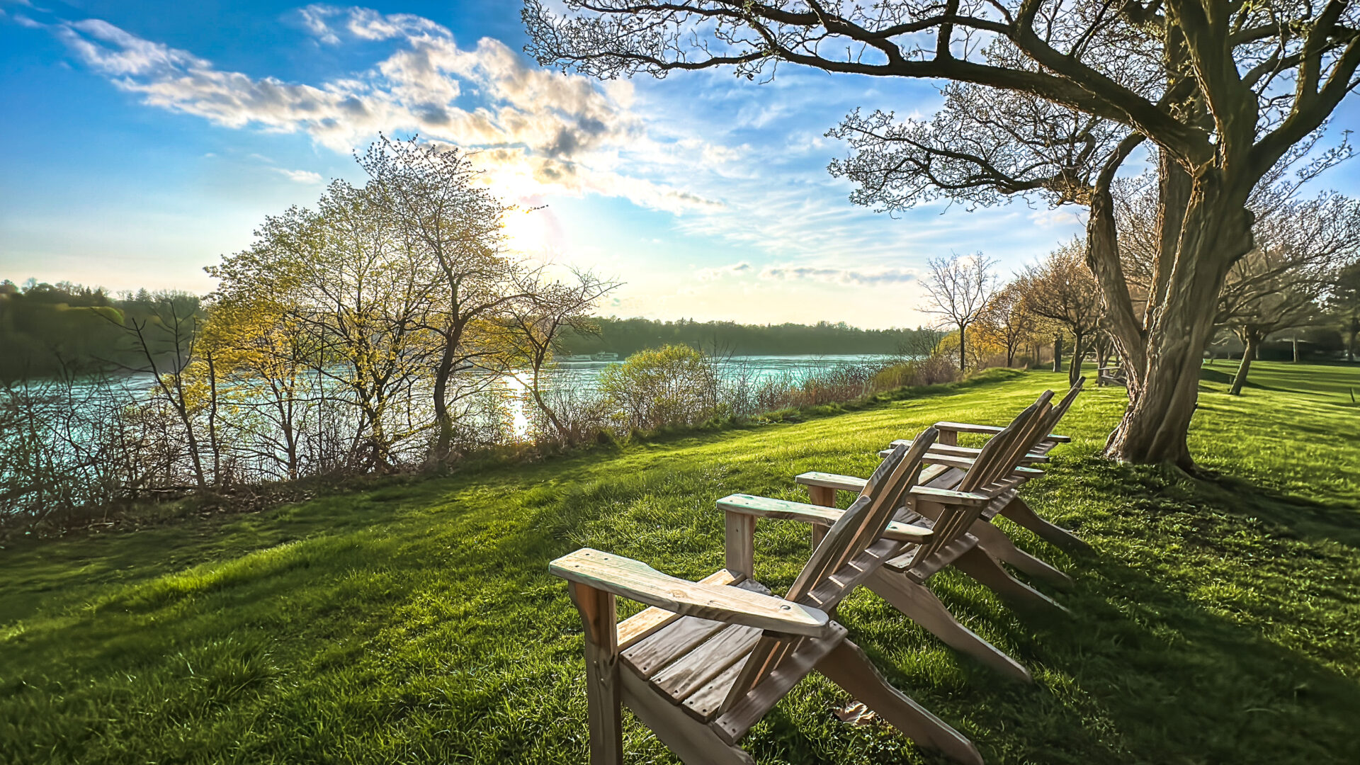 Sunset photo with dramatic skies, as the turquoise waters of the Niagara River flow in the background, while in the foreground, lush lawn bathed in golden light and three Adirondack chairs propped for prime viewing of the beaten path river scene