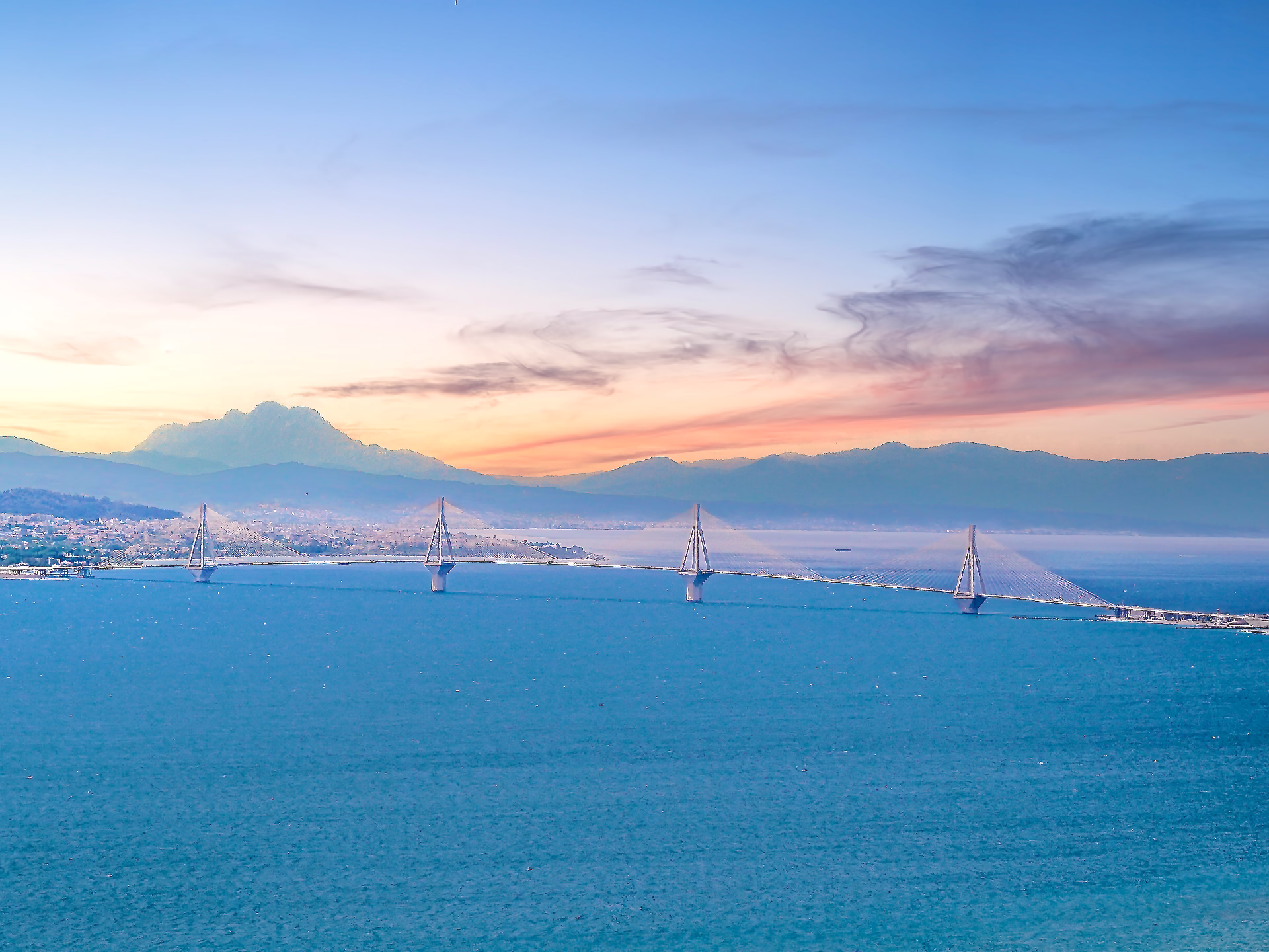 A view of the ocean with mountains in the background and the Rio-Antirrio bridge gleaming in a dramatic sunset