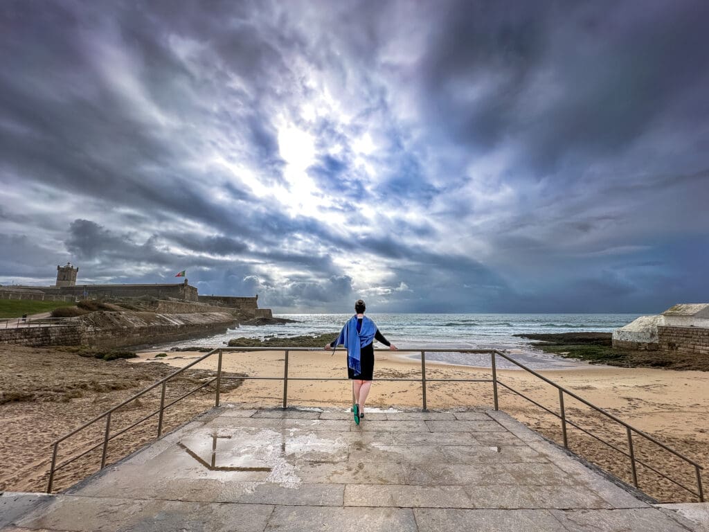 Person standing on a stone platform overlooking a dramatic cloudy sky, sandy beach, and ocean waves, with a historic fort featuring the Portuguese flag in the background, creating a peaceful coastal scene. Contemplating the new year ahead