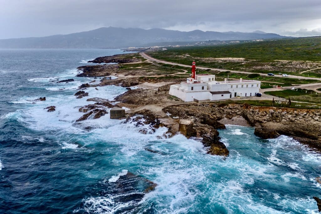 Aerial view of a red-and-white lighthouse perched on rugged cliffs, surrounded by the crashing waves of the Atlantic Ocean, with lush green hills and winding coastal roads in the background near Cascais, Portugal, perfect place to elect your new year resolution.