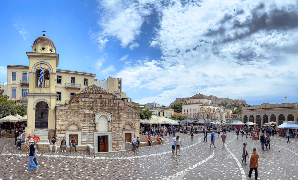 Panoramic view of Monastiraki Square in Athens, Greece, featuring a historic church and Tzistarakis Mosque, bustling crowds, and the Acropolis rising in the background under a partly cloudy sky. Monastiraki's flea market is full of hidden gems. Photo by Paul Kostadimas © spotlightsojourns.com