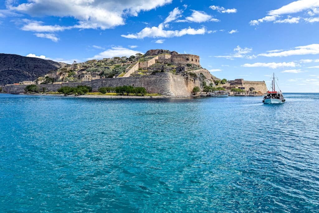 Scenic view of Spinalonga Island in Crete, Greece, featuring a historic Venetian fortress surrounded by turquoise waters, with a tourist boat approaching under a bright blue sky—popular cultural and historical travel destination. Photo by Paul Kostadimas © Spotlight Sojourns