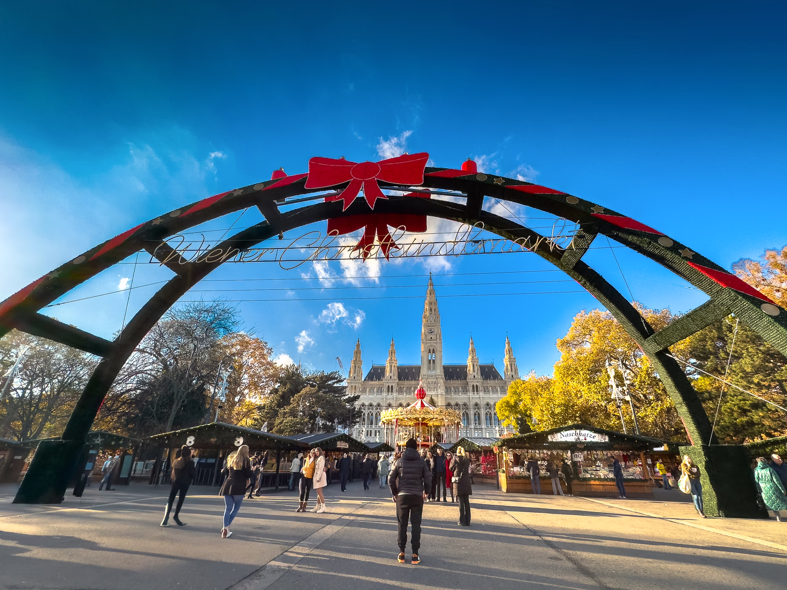 Exterior photo of a sunny winter's day infront of the decorated gates to the Rathausplatz Christkindlmarkt (Christmas market) at the Rathausplatz square.