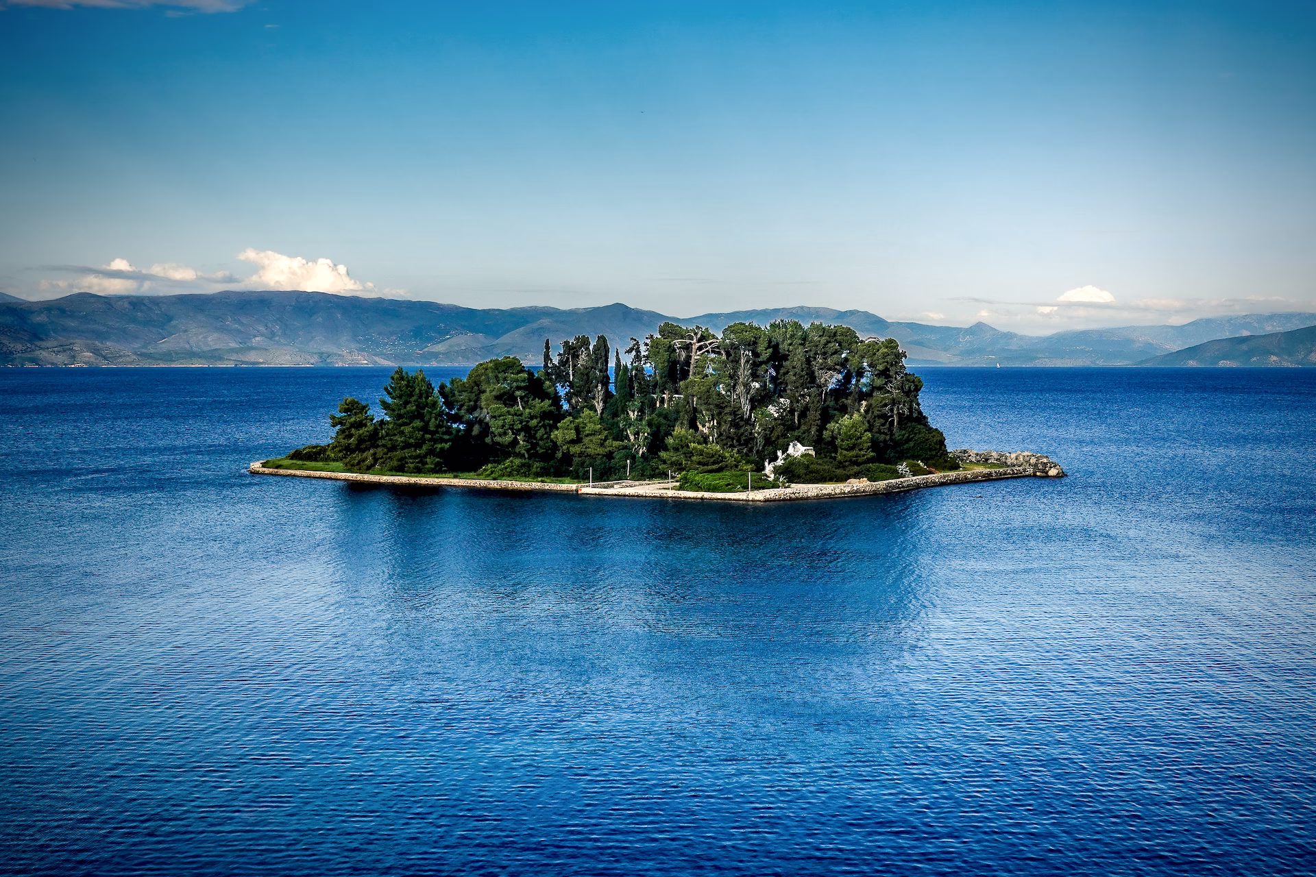 Pontikonisi (Mouse) Island - Corfu, Greece © Spotlight Sojourns Lush green island in the middle of a deep blue sea with mountains in the background. This is Pontikonisi The Legendary 'Mouse' Island in Corfu, Greece