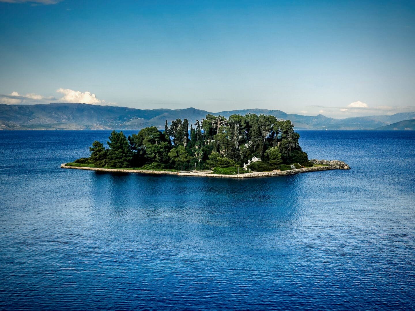 Pontikonisi (Mouse) Island - Corfu, Greece © Spotlight Sojourns Lush green island in the middle of a deep blue sea with mountains in the background. This is Pontikonisi The Legendary 'Mouse' Island in Corfu, Greece