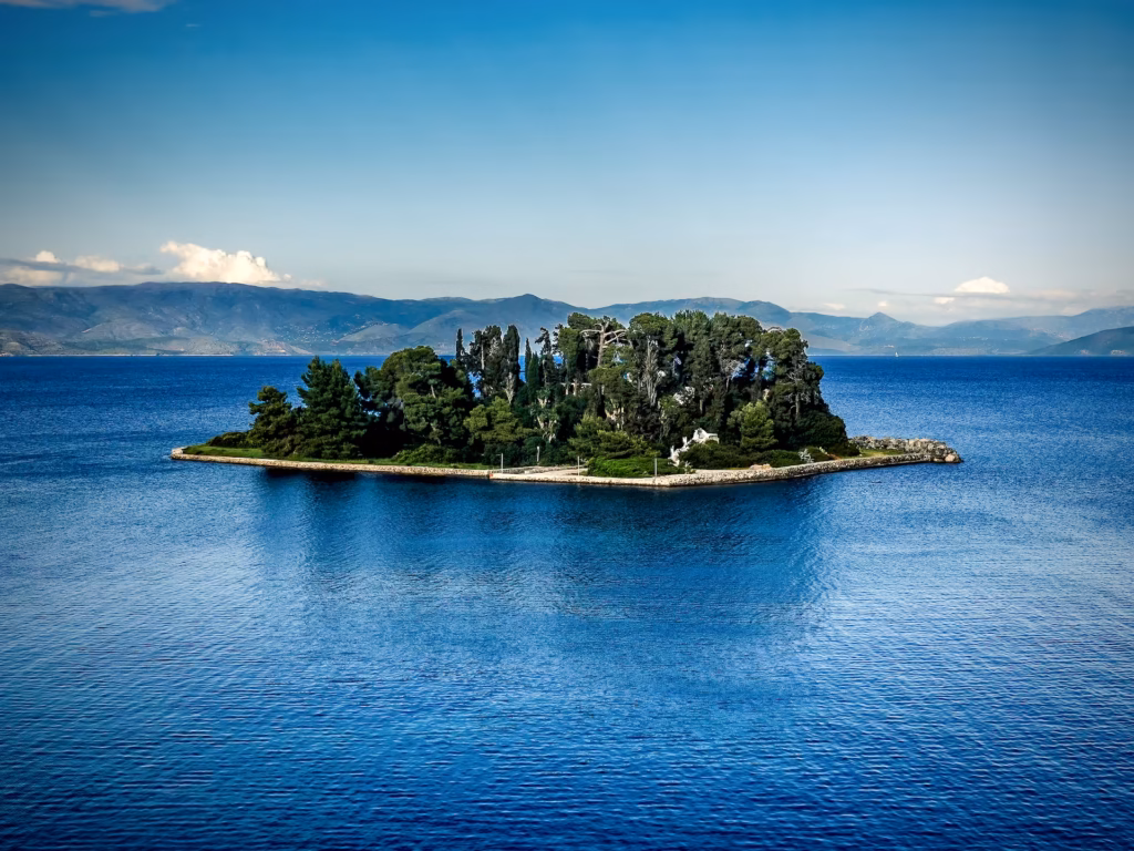 Lush green island in the middle of a deep blue sea with mountains in the background. This is Pontikonisi The Legendary 'Mouse' Island in Corfu, Greece