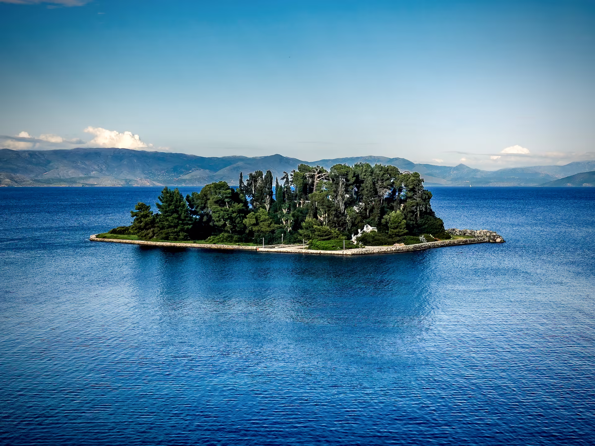 Lush green island in the middle of a deep blue sea with mountains in the background. This is Pontikonisi The Legendary 'Mouse' Island in Corfu, Greece