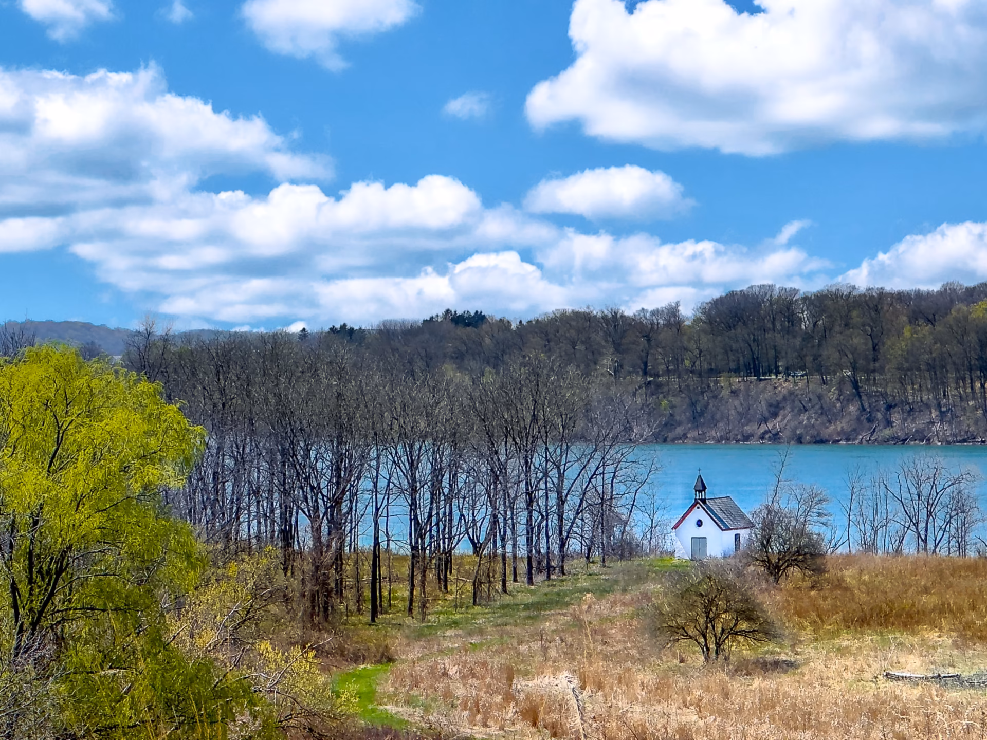 Stella Niagara preserve with trees starting to bloom, tall grass covers the foreground in front of a small white isolated church, the turquoise waters of the mighty Niagara River flow past, and a motor boat skims the water
