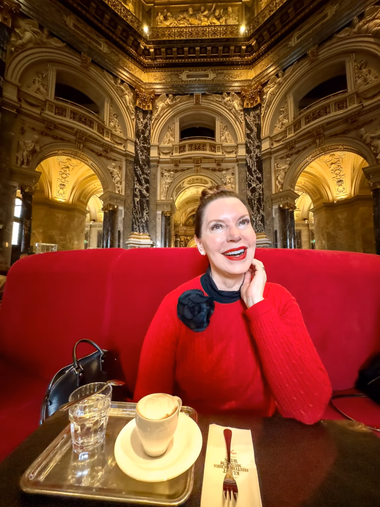 A woman sitting on top of a red couch next to an ornate ceiling. In the Kunst Historisches Museum Wien, there is a hidden gem of a coffee house