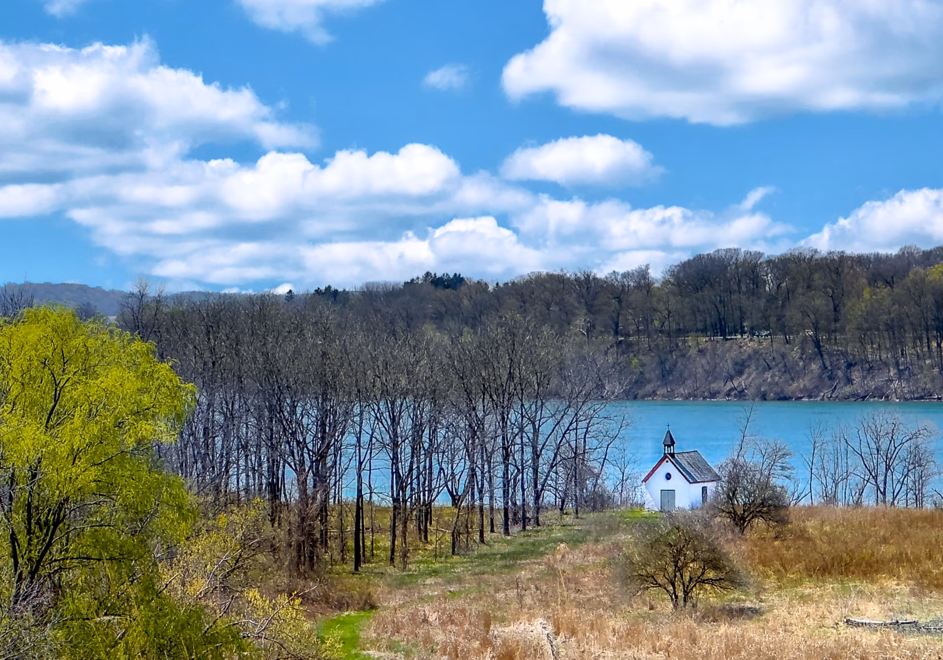 Stella Niagara preserve with trees starting to bloom, tall grass covers the foreground in front of a small white isolated church, the turquoise waters of the mighty Niagara River flow past, and a motor boat skims the water