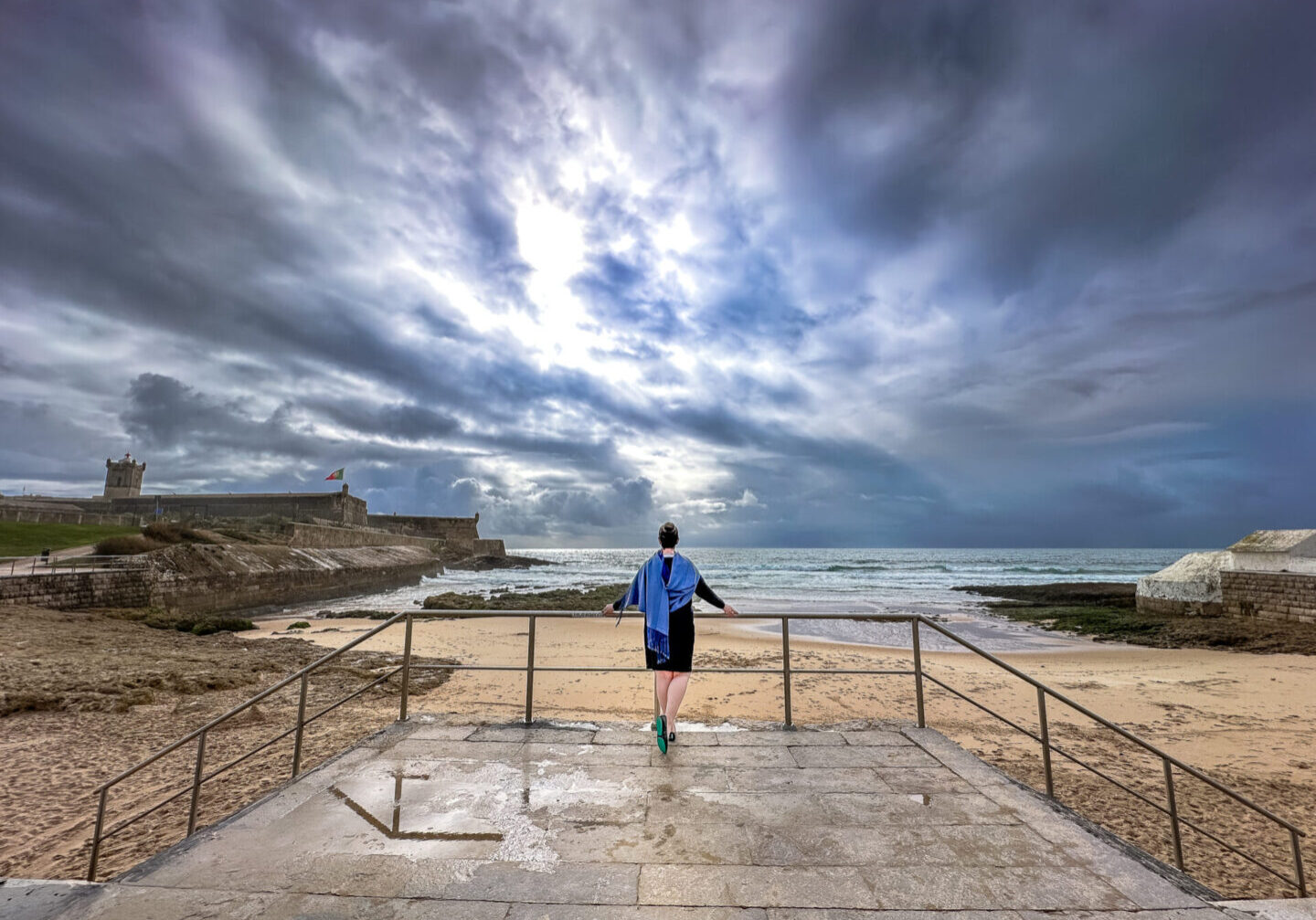 Person standing on a stone platform overlooking a dramatic cloudy sky, sandy beach, and ocean waves, with a historic fort featuring the Portuguese flag in the background, creating a peaceful coastal scene. Contemplating the new year ahead