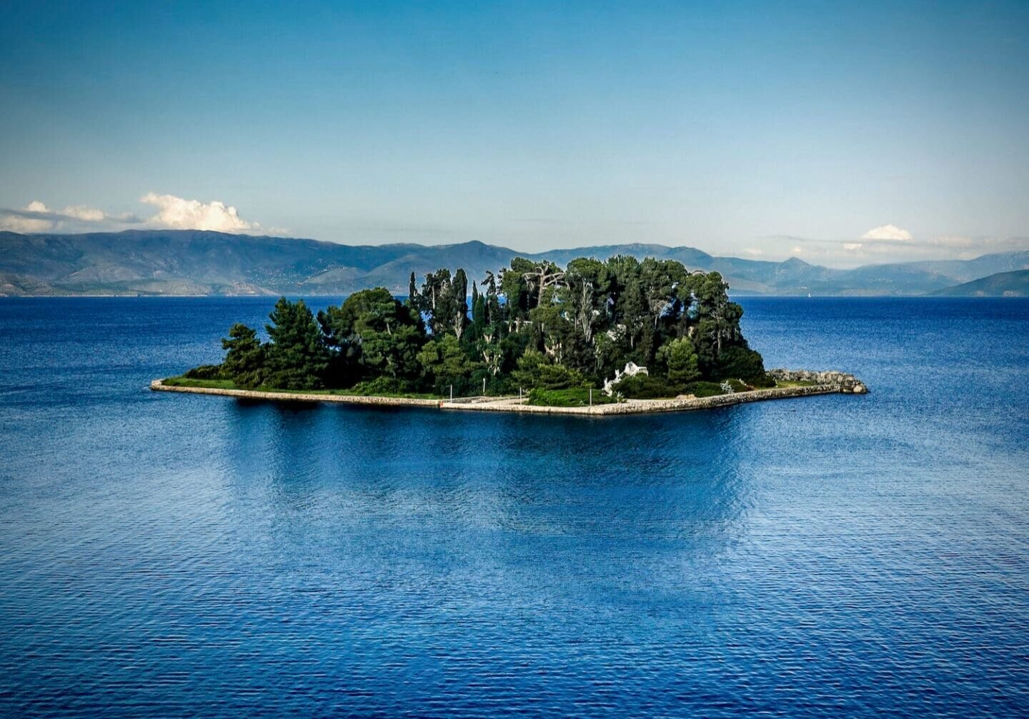 Lush green island in the middle of a deep blue sea with mountains in the background. This is Pontikonisi The Legendary 'Mouse' Island in Corfu, Greece