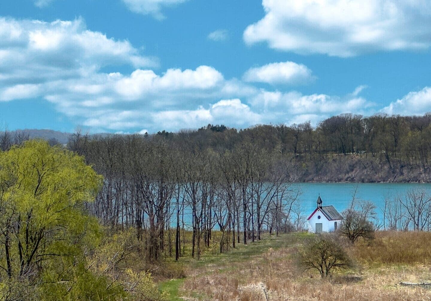 Stella Niagara preserve with trees starting to bloom, tall grass covers the foreground in front of a small white isolated church, the turquoise waters of the mighty Niagara River flow past, and a motor boat skims the water