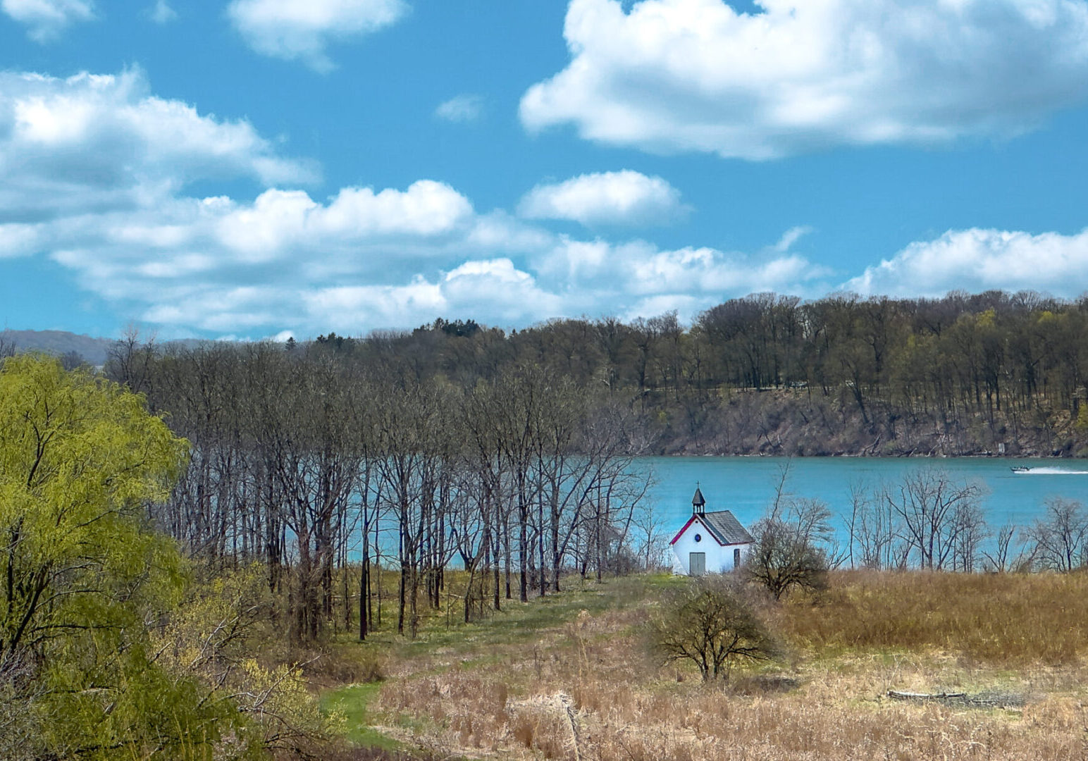 Stella Niagara preserve with trees starting to bloom, tall grass covers the foreground in front of a small white isolated church, the turquoise waters of the mighty Niagara River flow past, and a motor boat skims the water